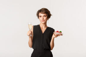 Stylish elegant woman attend party, holding glass of champagne and piece of cake, celebrating something over white background.